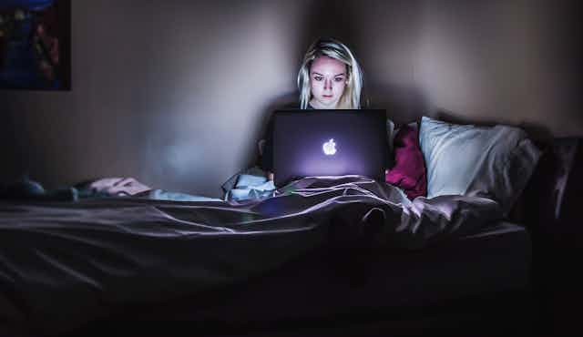 A woman with blonde hair sits in bed in the glow of her laptop.