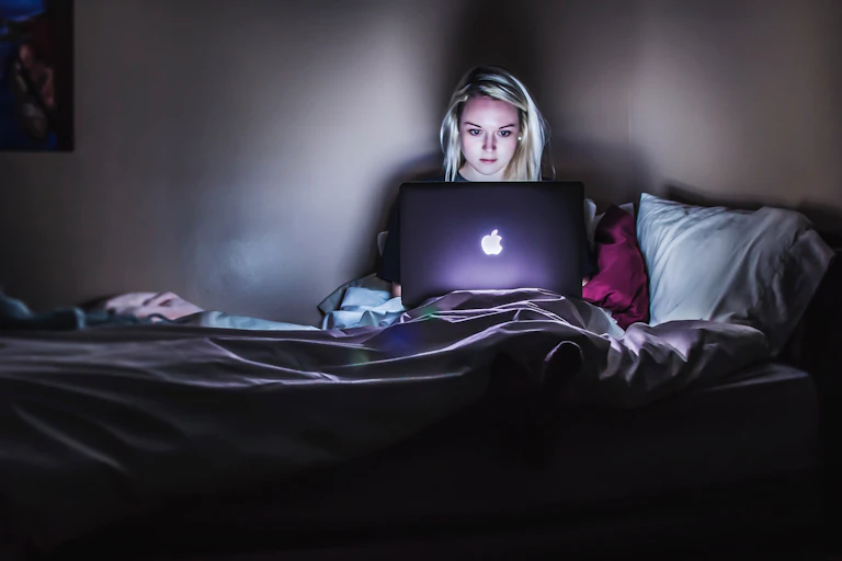 A woman with blonde hair sits in bed in the glow of her laptop.