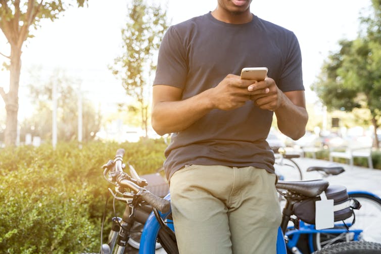 A man photographed from the neck down is seen looking at his phone while he leans against a bicycle outdoors.