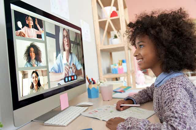 A young girl sits behind a computer monitor engaging with one older woman, and three other children her age
