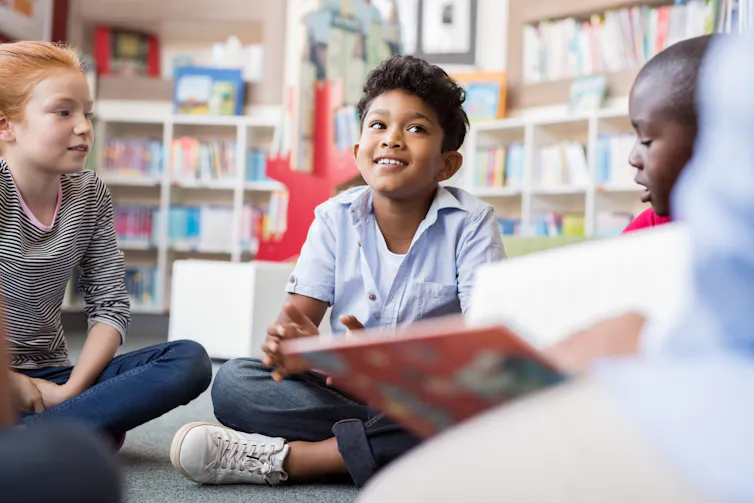 A group of kids sit cross-legged in a circle. Bookshelves are seen in the background.