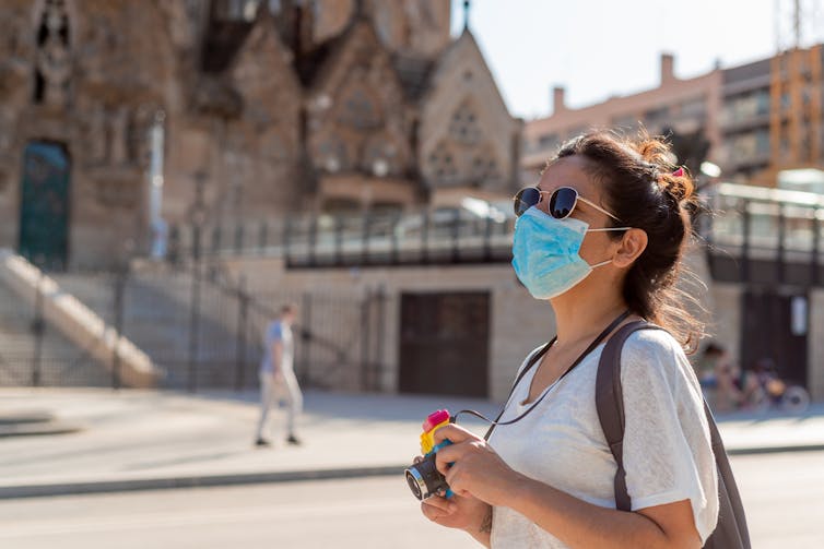 A young woman wearing a mask takes pictures in Barcelona, Spain. A building that looks like an old church is visible in the background.