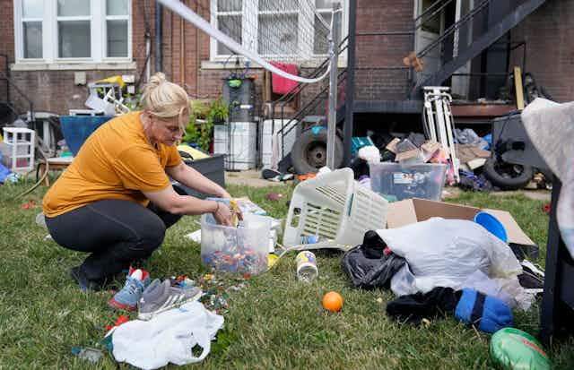 A woman kneels down on green grass as she gathers up her belongings that are strewn all over the ground in front of her former home