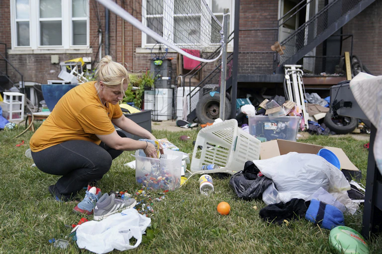 A woman kneels down on green grass as she gathers up her belongings that are strewn all over the ground in front of her former home