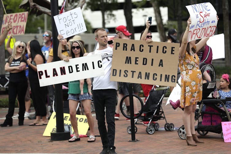 Protesters march carrying signs that read Plandemic and Practise Media Distancing.