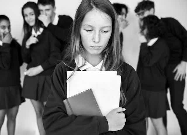 Black and white photo focusing on a sad looking girl in a school uniform, while her classmates laugh and whisper at her in the background