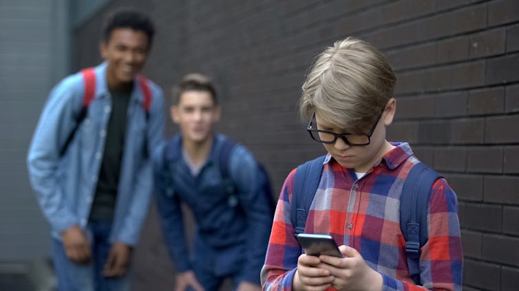 A young boy looks sadly at a mobile phone while two of his classmates laugh at him in the background.