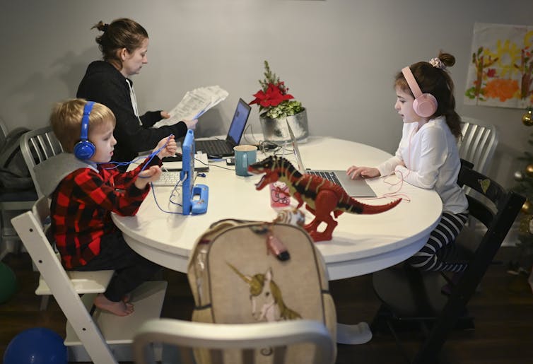 A mother sits at a crowded kitchen table in front of a laptop as two children do online schoolwork.