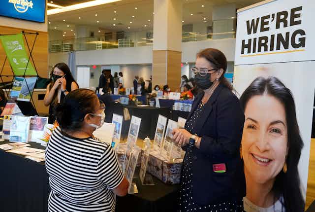 A female recruiter from Marriott hotels talks to a woman at a job fair at hard rock stadium in Miami Gardens Florida. a big sign says we're hiring and others talk on the phone in the background