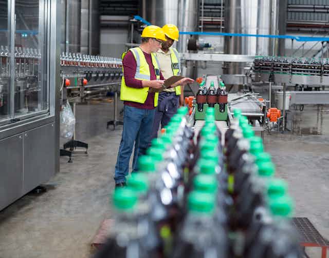 Two workers in hard hats and yellow vests inspecting a line of soft drink bottles on a conveyor belt