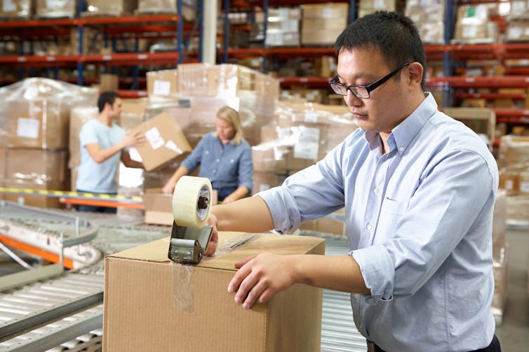 A warehouse with a worker in the foreground sealing a box with packing tape on a conveyor belt and two other workers in the background