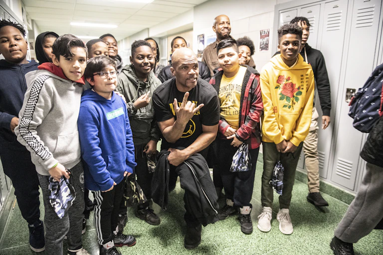 A rapper posses with a group of middle schools boys in a hallway.