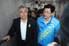 Two men, one in a suit jacket in the other a windbreaker jacket, stand shoulder to shoulder in a stairwell, photographers behind them