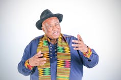 a man in a denim shirt with a colourful scarf and a hat smiles and gestures with his hands.