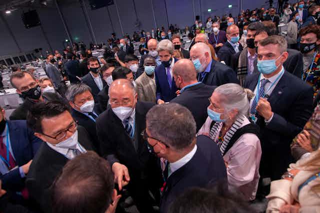 A large group of mostly men wearing suits and face masks talk to each other.