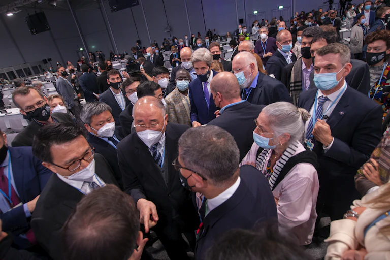 A large group of mostly men wearing suits and face masks talk to each other.