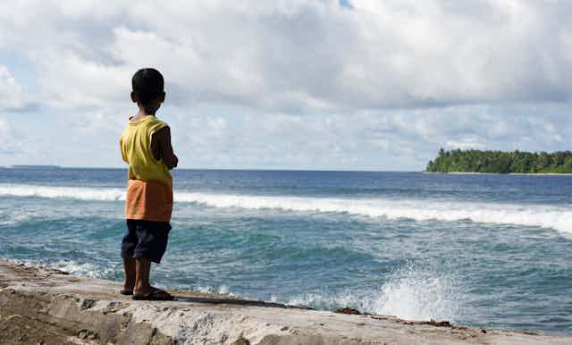 A boy stands on a sea wall on a tropical island overlooking the tide.