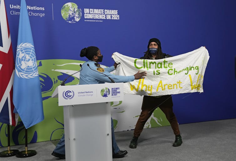 woman at lectern looks at woman holding banner