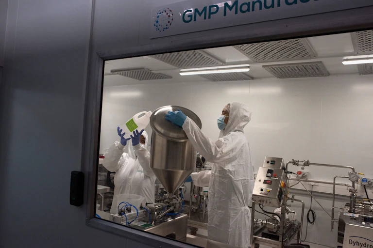 Two people in white hazmat suits are seen through a laboratory window handling a big steel container. They are surrounded by laboratory equipment