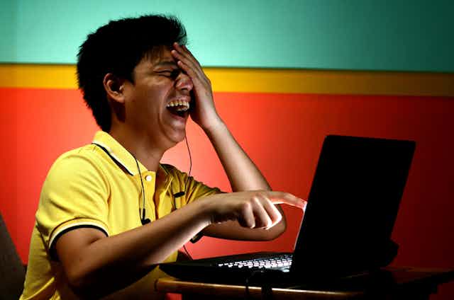 A young man laughs hysterically while pointing at something on a computer screen.
