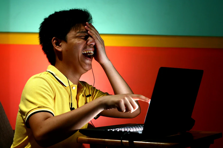 A young man laughs hysterically while pointing at something on a computer screen.