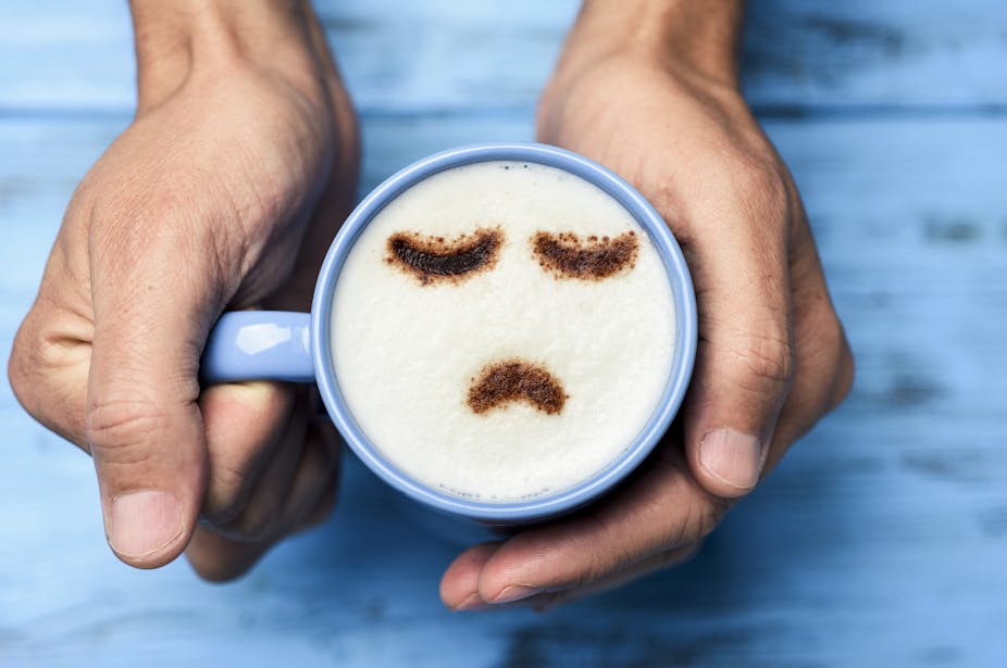 A person holding a mug of coffee in their hands. On the foam is a frowning face made out of chocolate powder.