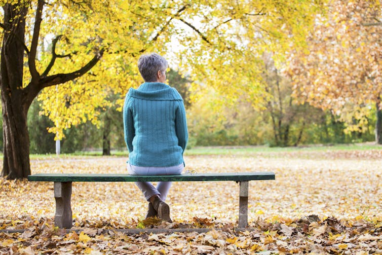 An older woman sitting on a bench in a park looking at the autumn leaves.