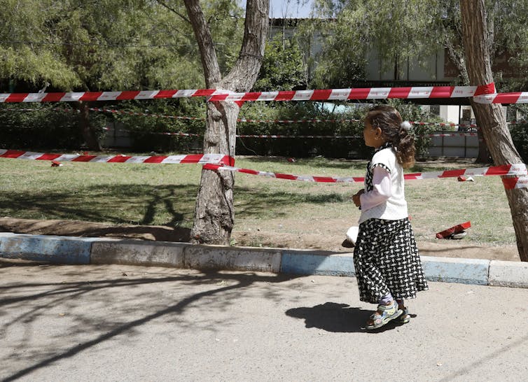 A young girl walks past a garden that has been roped off with red and white tape.