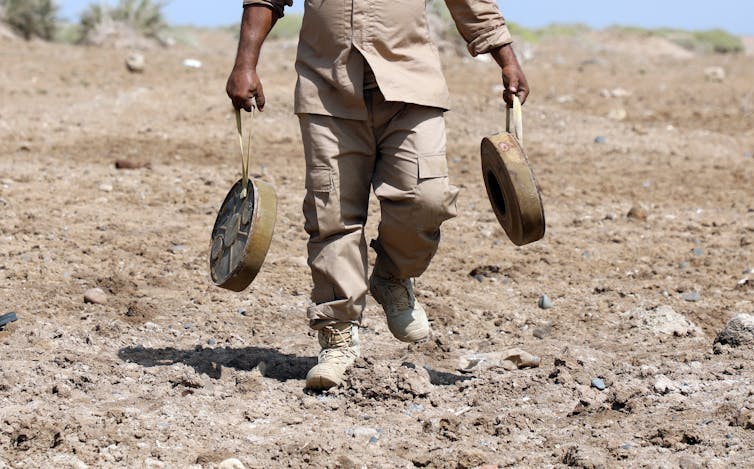 Man carrying two anti-personnel mines across bare soil.
