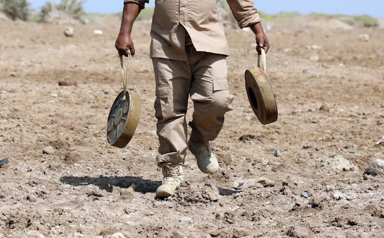 Man carrying two anti-personnel mines across bare soil.