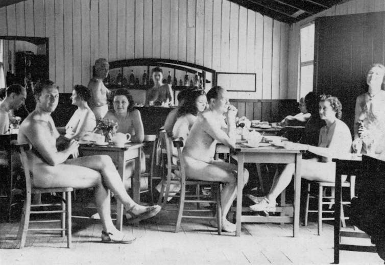 A group of men and women sit at tables, naked apart from their shoes