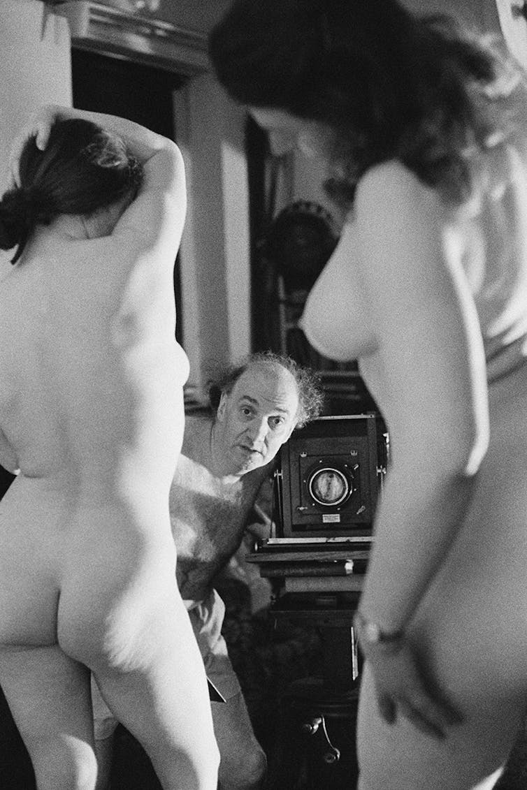 A middle-aged man with a camera peers between the bodies of two young nude women.