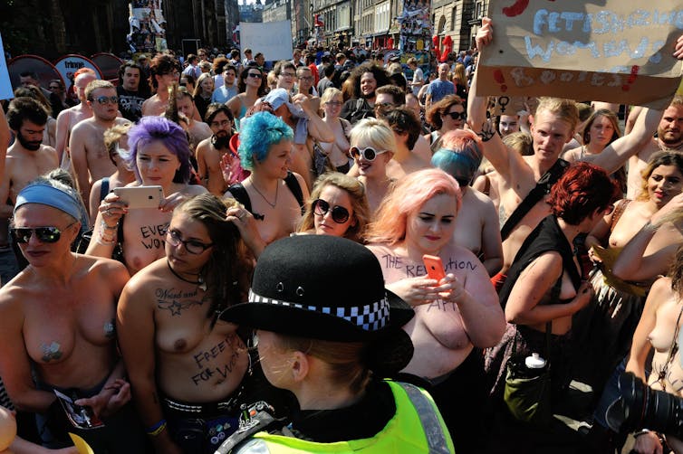 A crowd of topless women holding placards stand in front of a policewoman.