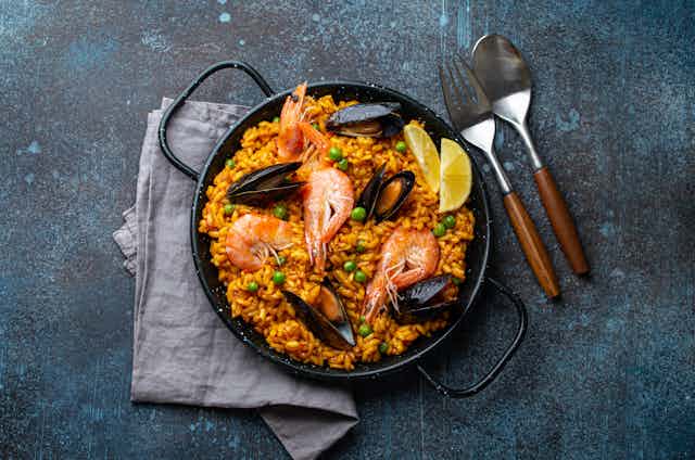 Overhead view of a dish of bright orange paella with prawns and mussels on a blue concrete table.