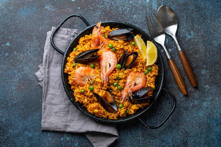 Overhead view of a dish of bright orange paella with prawns and mussels on a blue concrete table.