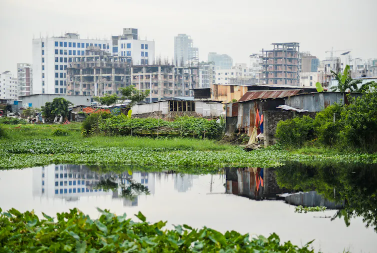 In the foreground, a body of water reaches close to precarious housing structures. In the background, highrise buildings tower.