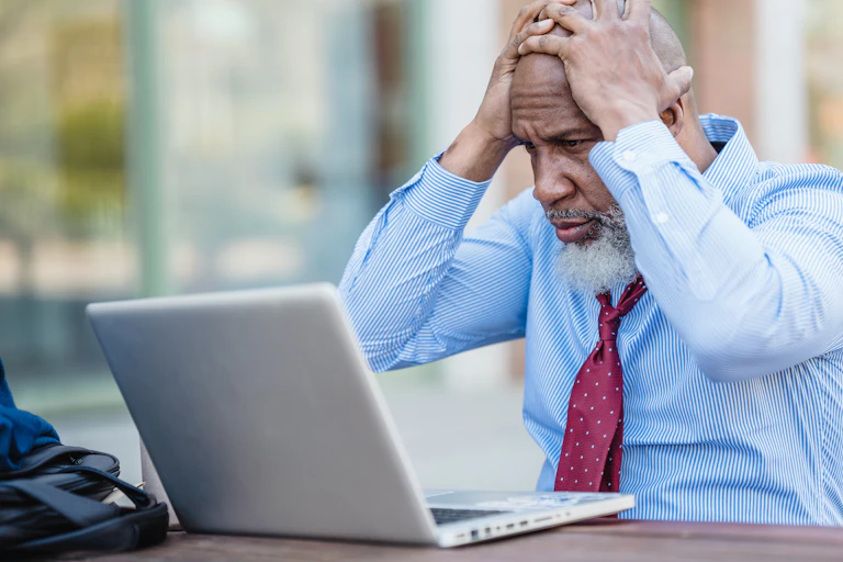 A bearded man in a blue shirt and red tie looks at a laptop with his hands on his head.