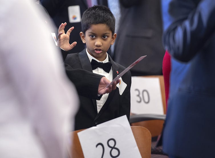 A young boy in a tuxedo raises his right arm.