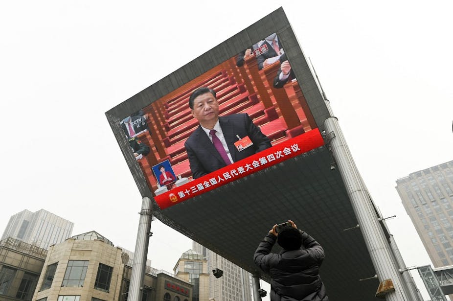 Un homme de dos prend en photo un écran géant qui retransmet Xi Jinping en costume sur fond rouge.