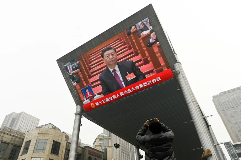 Un homme de dos prend en photo un écran géant qui retransmet Xi Jinping en costume sur fond rouge.