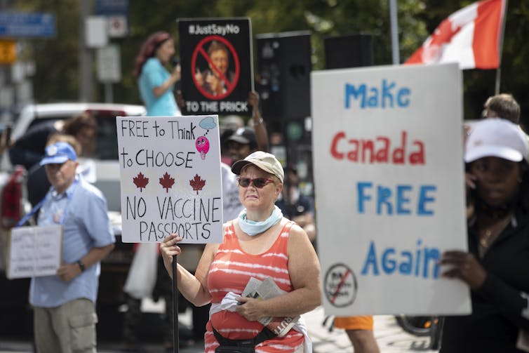 A group of protesters, including a woman in an orange-and-white tank top, blue scarf and baseball hat, hold signs, one of which says Make Canada Free Again.