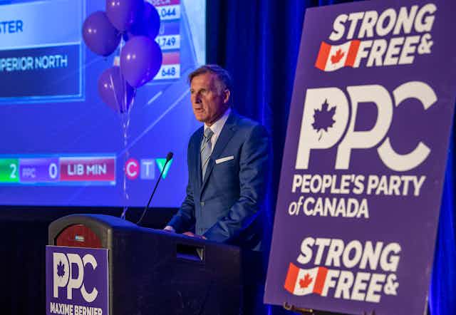 A man in a blue suit speaks to supporters from a podium with a PPC sign next to him. 