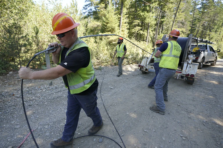 Four people wearing reflective safety vests and hardhats pull a black cable from a large spool on a trailer behind a pickup truck on a rural road