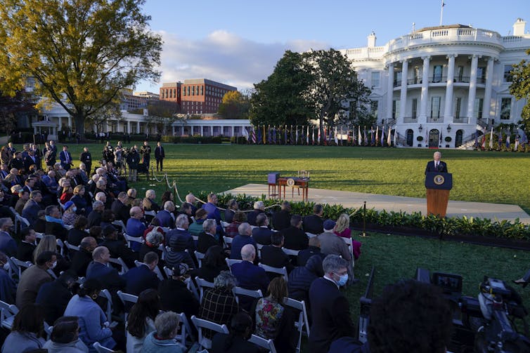With the White House in the distance, President Biden speaks at a lectern before a crowd of people