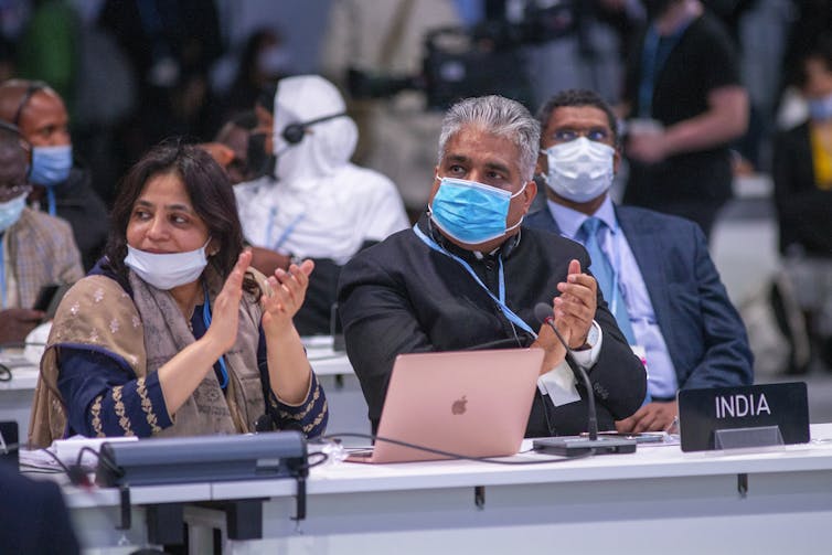 Two delegates wearing face masks sit at a white desk with a sign reading 'India' on it.