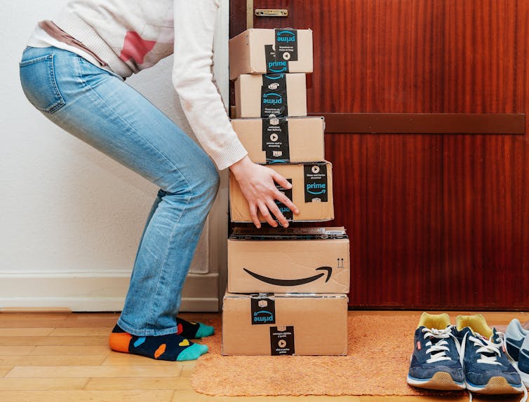 A person bending down to lift a large stack of Amazon boxes in their home