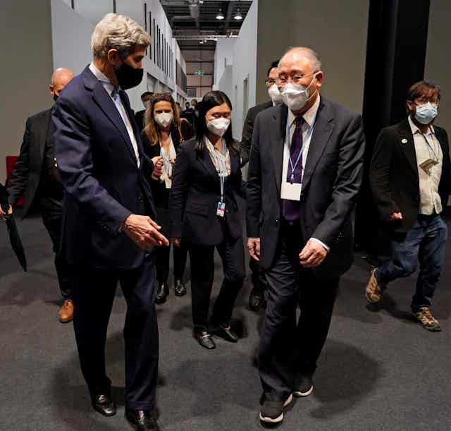 Two men in suits walk down a hallway followed by a group of people and journalists.