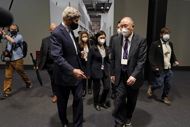 Two men in suits walk down a hallway followed by a group of people and journalists.
