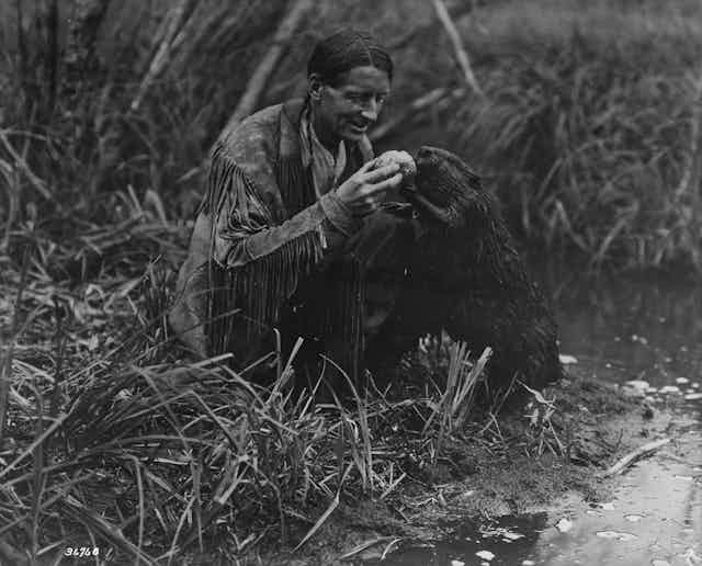 A black and white image of a man with a leather fringe jacket, feeding a beaver a jelly roll.