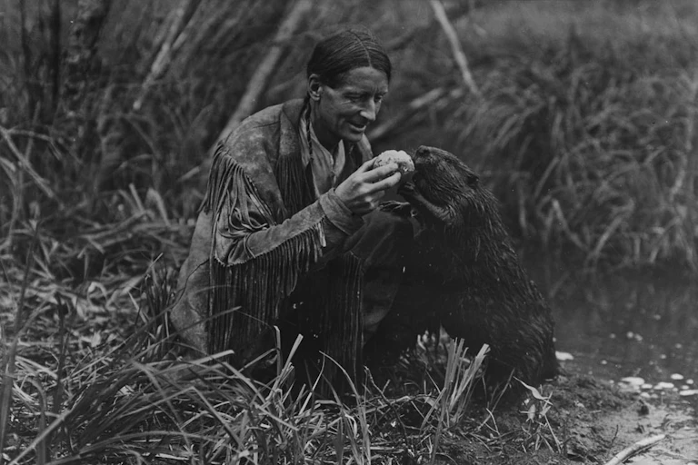A black and white image of a man with a leather fringe jacket, feeding a beaver a jelly roll.